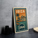 A vintage-style metal sign with the text "IBiza" and "SPAIN" printed on it, depicting a tropical island scene with palm trees and buildings.