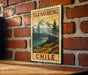 A vintage-style postage stamp depicting a scenic landscape with mountains, a lake, and a path, hanging on a brick wall.