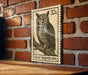 A postage stamp featuring a great horned owl is displayed on a brick wall.