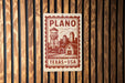 A sign with the text "PLANO TEXAS USA" and an illustration of a water tower, barn, and field, mounted on a wooden wall.