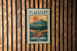 A vintage-style metal sign with a mountain landscape and a sun in the sky, mounted on a wooden wall.