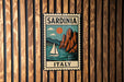 A vintage-style metal sign with the text "SARDINIA ITALY" and an illustration of a sailboat on a body of water, mounted on a wooden wall.