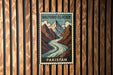 A vintage postage stamp featuring a mountain landscape with a river, mounted on a wooden wall.