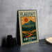 A metal sign with a vintage-style illustration of a mountain landscape and a sun, along with the text "FLAGSTAFF ARIZONA USA".