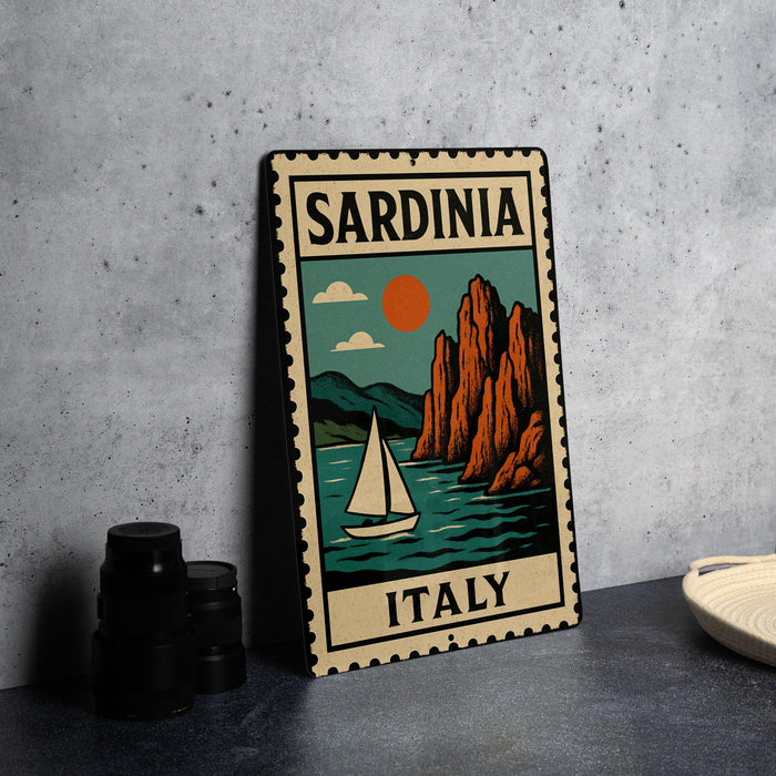A vintage-style metal sign with the text "SARDINIA ITALY" and an illustration of a sailboat on a body of water, with mountains in the background.