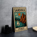 A vintage-style metal sign with the text "SARDINIA ITALY" and an illustration of a sailboat on a body of water, with mountains in the background.