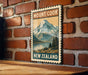 A vintage-style poster of Mount Cook, a famous mountain range in New Zealand, is displayed on a brick wall.