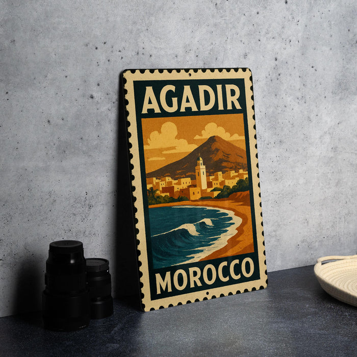 A vintage-style metal sign with the text "AGADIR MOROCCO" and an illustration of a coastal city with mountains in the background.