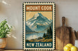 A vintage-style metal sign with a mountain landscape and a lake, advertising "Mount Cook New Zealand". The sign is displayed on a shelf next to a wooden cutting board and some kitchen utensils.