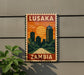 A vintage-style sign that reads "LUSAKA ZAMBIA" with an illustration of a cityscape and a sunset.