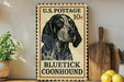 A vintage-style U.S. postage stamp featuring a blue and white spotted dog, with the text "BLUE TICK COONHOUND" printed on it. The stamp is placed on a wooden surface, with a potted plant, a wooden cutting board, and some lemons nearby.