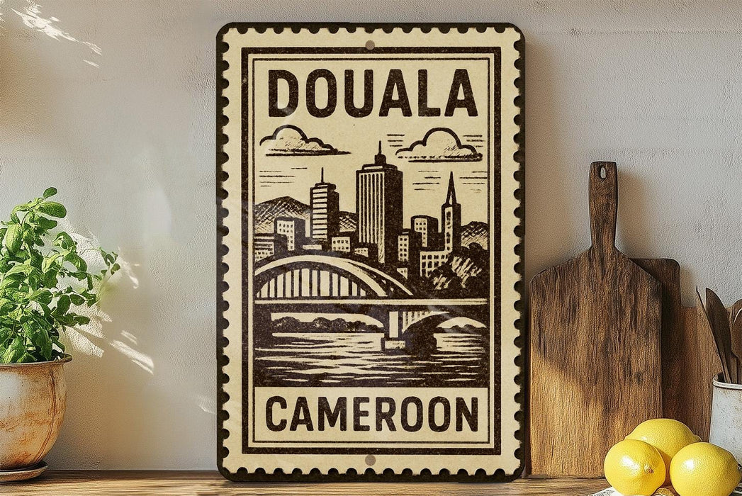 a decorative metal sign with the text "DOUALA CAMERON" and an illustration of a cityscape with a bridge. The sign is placed on a shelf next to a potted plant and a wooden cutting board.