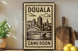 a decorative metal sign with the text "DOUALA CAMERON" and an illustration of a cityscape with a bridge. The sign is placed on a shelf next to a potted plant and a wooden cutting board.