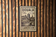 a stamp-like sign with the text "DOUALA CAMERON" and a cityscape illustration, mounted on a wooden wall.