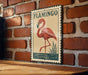 A vintage-style metal sign featuring a pink flamingo standing on one leg in a pond, mounted on a brick wall.