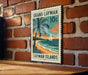 A vintage-style postage stamp depicting a tropical beach scene with a sailboat, palm trees, and a sunset over the ocean, mounted on a brick wall.