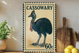 A vintage-style postage stamp featuring a black ostrich, with the text "CASSOWARY" and "60c" printed on it. The stamp is displayed on a shelf next to a wooden cutting board and a potted plant.
