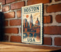 A vintage-style poster of Boston, Massachusetts, featuring the city's iconic landmarks and skyline, is displayed on a brick wall.
