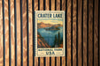 A vintage-style metal sign for Crater Lake National Park in the United States, featuring a scenic landscape with mountains, a lake, and pine trees.