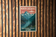 A vintage-style postage stamp depicting a scenic landscape with mountains, a river, and pine trees, mounted on a wooden wall.