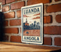 A metal sign with the text "LUANDA ANGOLA" and an illustration of a cityscape with a river and a boat.