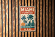 A vintage-style stamp or sign with the text "MIAMI FLORIDA" and a graphic of palm trees and a sunset, mounted on a wooden wall.