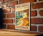 A vintage-style poster of the Twelve Apostles, a famous limestone stacks in Australia, is displayed on a brick wall.