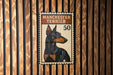 A Manchester Terrier dog is depicted on a postage stamp-like sign, which is mounted on a wooden wall.