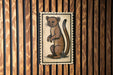 a wooden wall with a framed illustration of a brown animal, possibly a rodent or a small mammal, mounted on it.