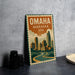 A vintage-style metal sign with the text "OMAHA NEBRASKA USA" and an illustration of a cityscape, placed on a gray countertop next to some black camera lenses.