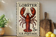 A vintage-style stamp featuring a lobster, with the text "LOBSTER 5c U.S. POSTAGE" printed on it. The stamp is displayed on a shelf next to a wooden cutting board and some kitchen utensils.