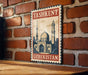 A vintage-style metal sign with the text "TASHKENT UZBEKISTAN" and an illustration of a mosque and a tower, mounted on a brick wall.