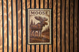 A framed image of a moose is displayed on a wooden wall.