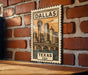A framed illustration of a cityscape with a bridge and a tower, mounted on a brick wall.