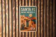 A vintage-style poster of Santa Fe, New Mexico, featuring a desert landscape with a cactus and mountains in the background.