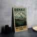 A metal sign with a mountain landscape and pine trees, featuring the text "DENALI NATIONAL PARK USA".