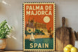 A vintage-style metal sign with the text "PALMA DE MAJORCA" and "SPAIN" printed on it, featuring an illustration of a castle and a sailboat. The sign is displayed on a shelf next to a wooden cutting board and a potted plant.
