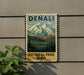 A vintage-style metal sign with the text "DENALI NATIONAL PARK USA" displayed on a wall, featuring a mountain landscape with pine trees.