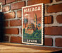 A vintage-style metal sign with the text "MALAGA" and "SPAIN" displayed on it, featuring an illustration of a building and palm trees.