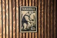 A stamp depicting an animal, likely an ant, with the text "TAMANDU" printed on it. The stamp is mounted on a wooden wall with vertical slats.