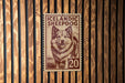 A brown and white sheepdog is depicted on a brown and white postage stamp, which is affixed to a wooden wall.