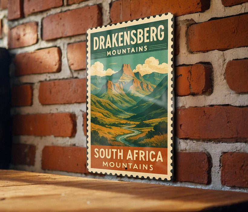 A vintage postage stamp featuring a mountain landscape with the text "DRAKENSBERG MOUNTAINS SOUTH AFRICA" is displayed on a brick wall.