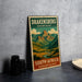 A metal poster with a mountain landscape and text, placed on a gray surface next to some black camera lenses.