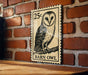A wooden table with a wooden post on it, and a vintage-style 25c postage stamp featuring an illustration of a barn owl on it, mounted on a brick wall.
