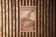 A red and white postage stamp from Russia with a mountain and ocean illustration, mounted on a wooden wall.