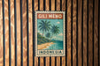A vintage postage stamp featuring a tropical beach scene with palm trees and a blue ocean, mounted on a wooden wall.