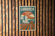 A vintage-style postage stamp from Texas, featuring a cityscape and a barn, is displayed on a wooden wall.