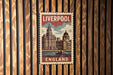 A metal sign with the text "LIVERPOOL ENGLAND" and an illustration of a city skyline, mounted on a wooden wall.