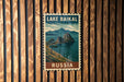 A vintage-style postage stamp depicting Lake Baikal, the world's largest cave system, with mountains and a lake in the background. The stamp is displayed on a wooden wall.