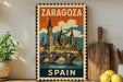 A vintage-style metal sign with the text "ZARAGOZA SPAIN" and an illustration of a castle or cathedral, placed on a wooden surface next to a potted plant and a wooden cutting board.